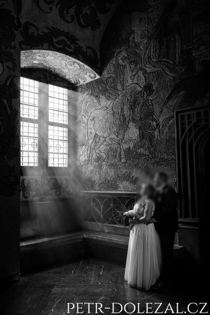 Černobílá fotografie nevěsty a ženicha pod oknem skrze které prosvítají paprsky slunečního světla