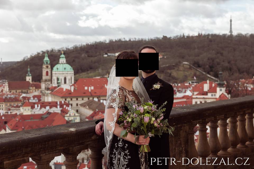 Portrétní fotografie ženicha a nevěsty na venkovní terase se zdobným zábradlím, v pozadí panorama Prahy s dominantní kupolí malostranského kostela sv. Mikuláše