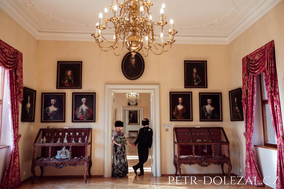 Ženich s nevěstou v salonku Paláce Lobkowicz, velký zdobný lustr, na stěnách historické portrétní malby, historický nábytek.