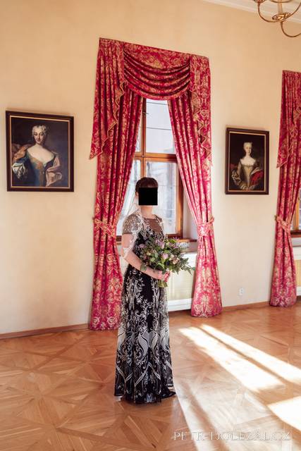 Portrét celé postavy nevěsty před oknem salonku, vedle okna jsou na stěně obrazy s portréty žen