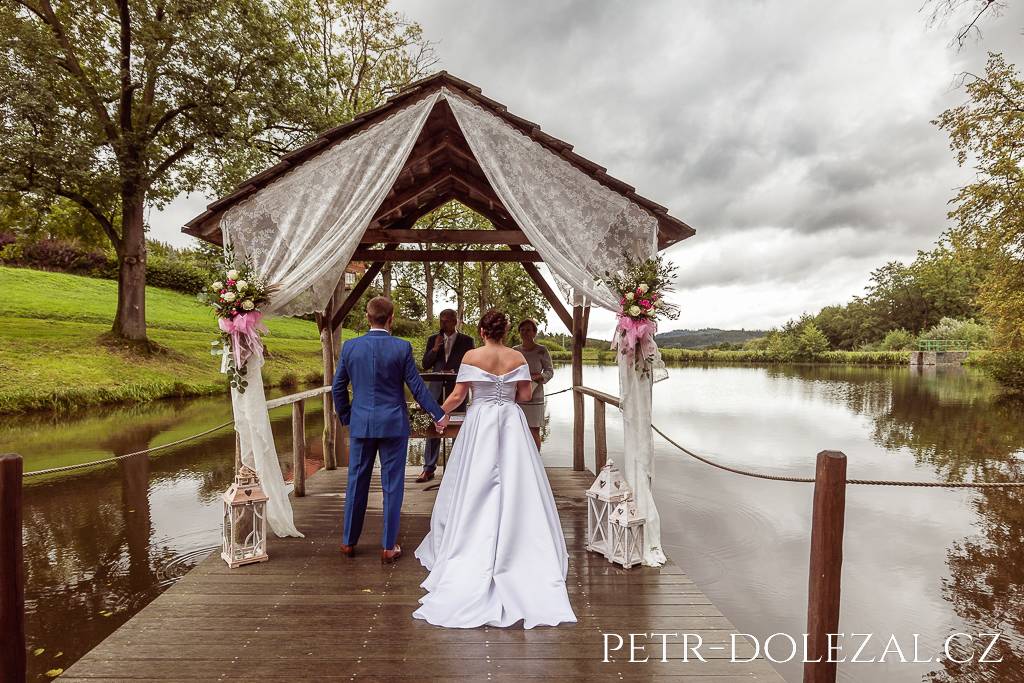 Ženich s nevěstou vyfocení zezadu v průběhu svatebního obřadu na zastřešeném molu u rybníka