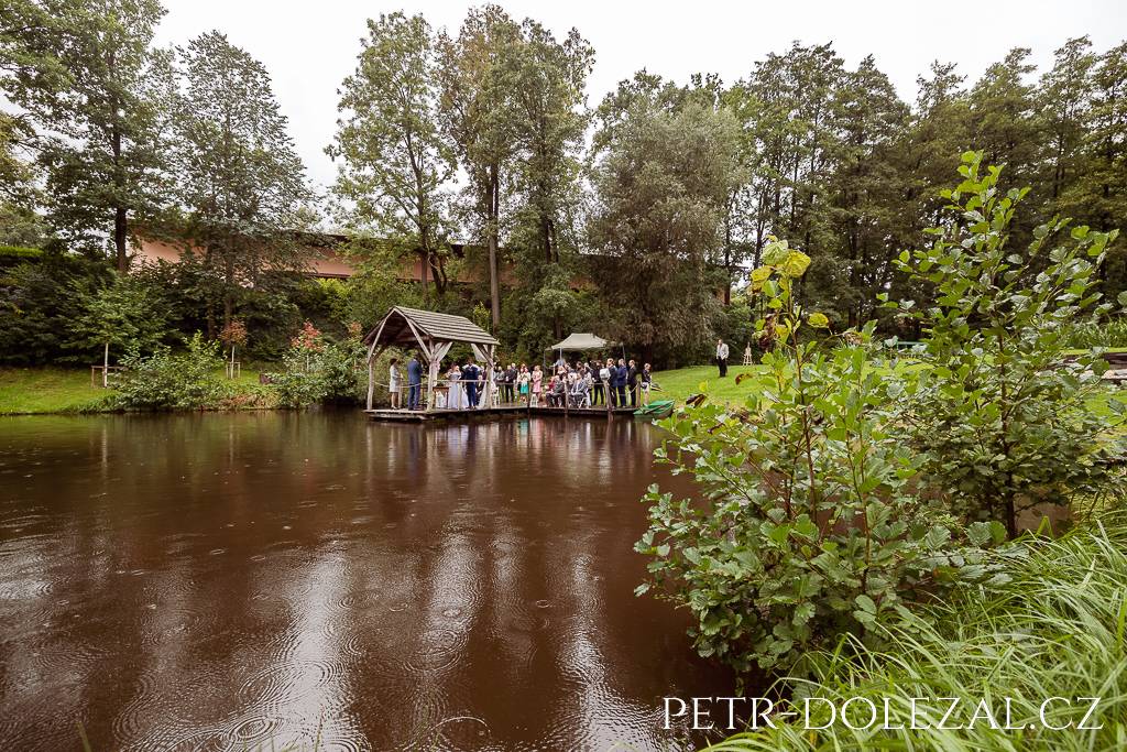 Svatební obřad na kraji rybníka vyfocený z větší vzdálenosti s důrazem na kruhy v hladině rybníka z dešťových kapek