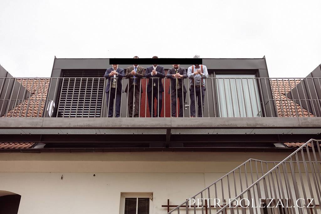 Fotografie ženicha a kamarádů opírajících se o zábradlí ve vyvýšeném patře, fotografováno ze země z podhledu
