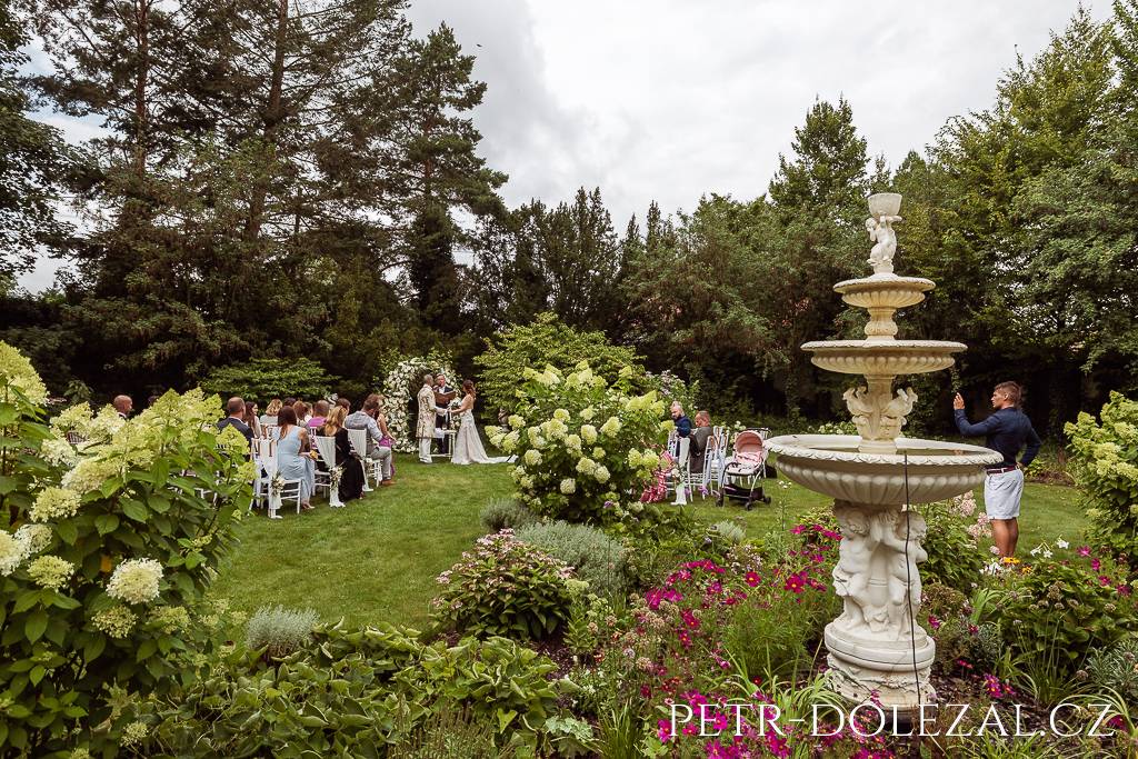 Pohled na ženicha s nevěstou a další svatebčany přes květinový záhon a kašnu při probíhajícím svatebním obřadu