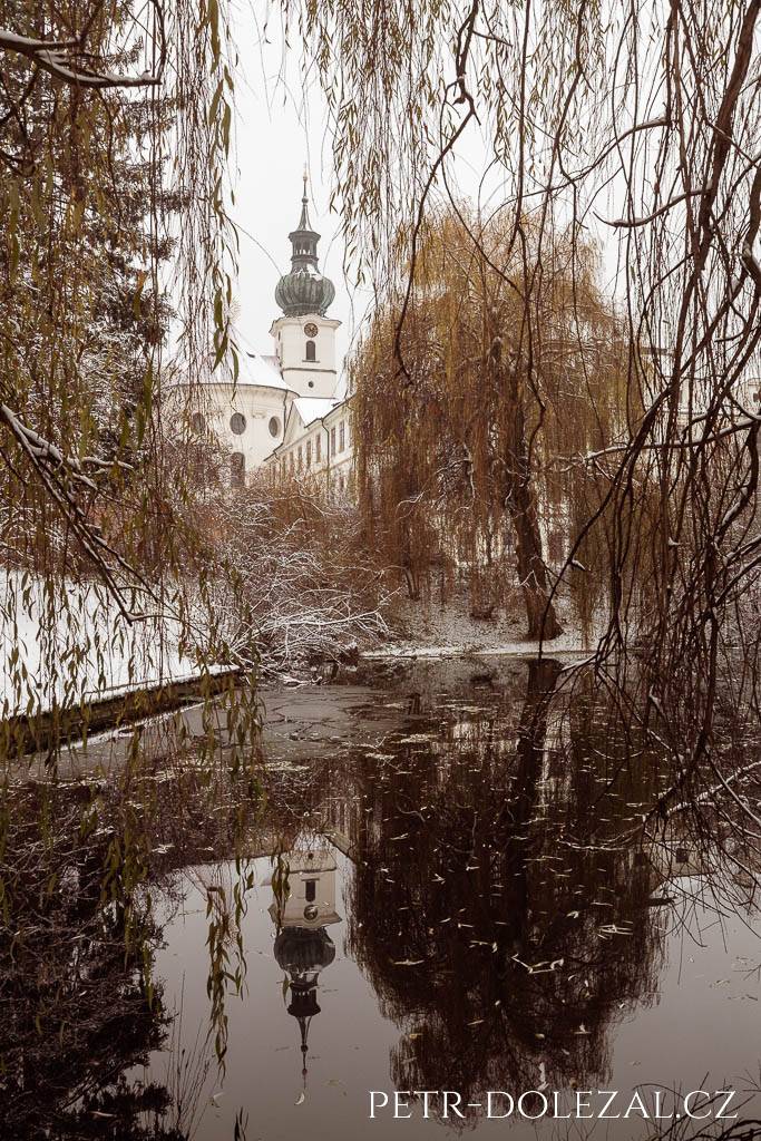 Kostel sv. Markéty zrcadlící se v rybníku Markéta II. v zimě se sněhem, průhled přes větve vrby