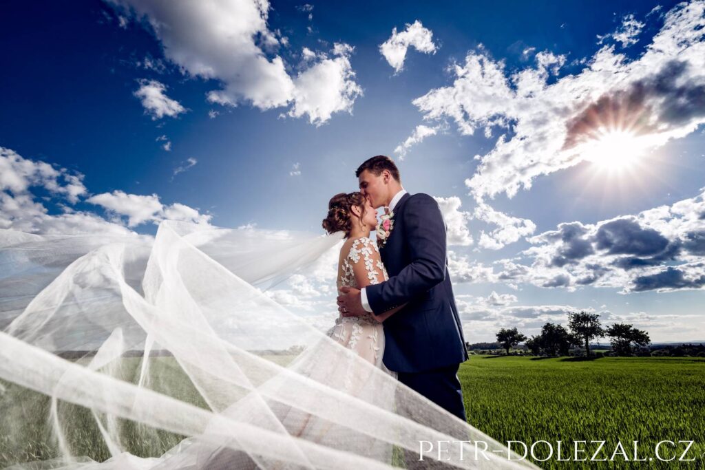 the bride and groom embracing, the bride with her veil blowing in the wind, the field and the sun in the background