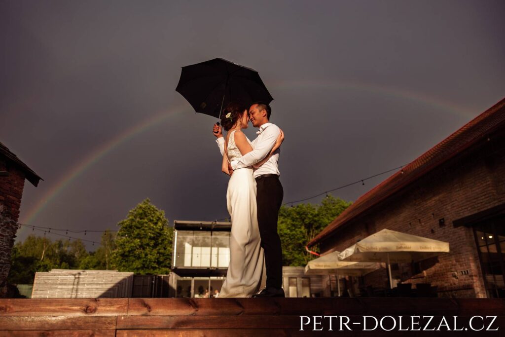 Bride and groom embrace with umbrella and rainbow above their heads