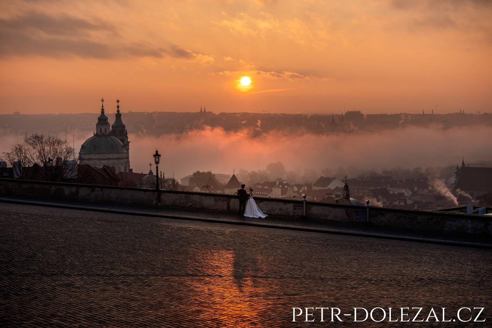 Groom and bride look out over the panorama of Prague rooftops at sunrise, foggy haze over Prague