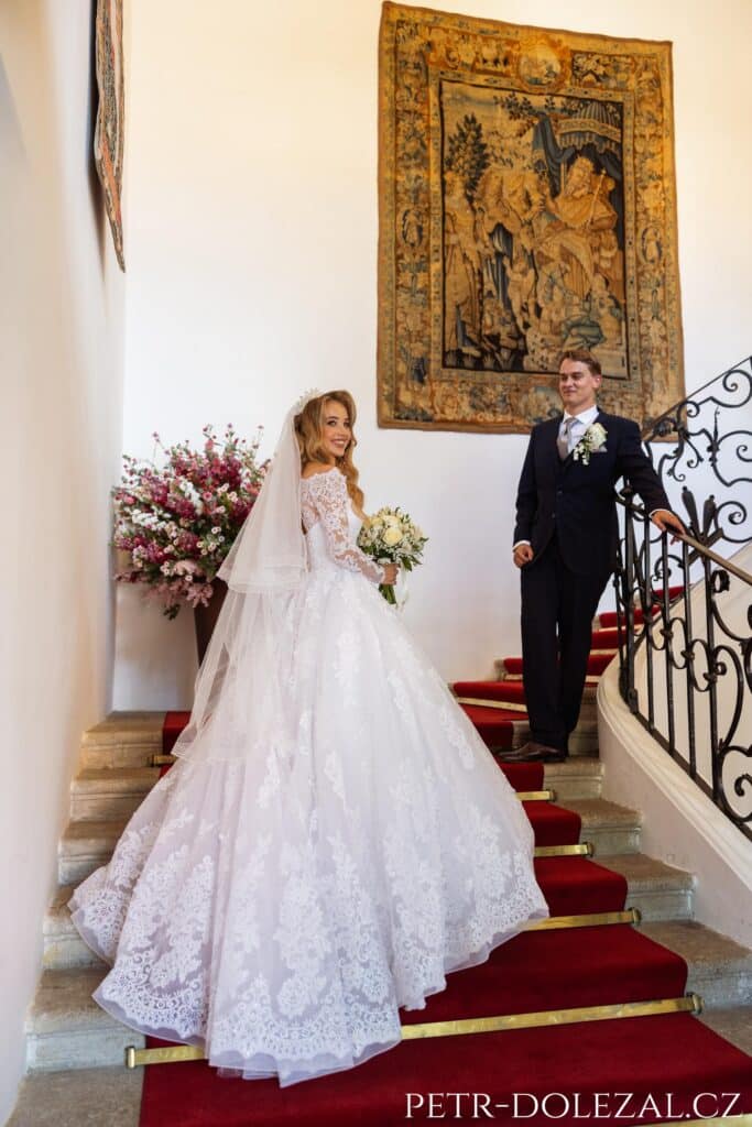 Nevěsta a ženich na schodech uvnitř zámku. Schodiště je potažené červeným kobercem. Nevěsta stojí více dole, zády otočená tak, aby byly vidět zezadu její šaty splývající dolů ze schodů.