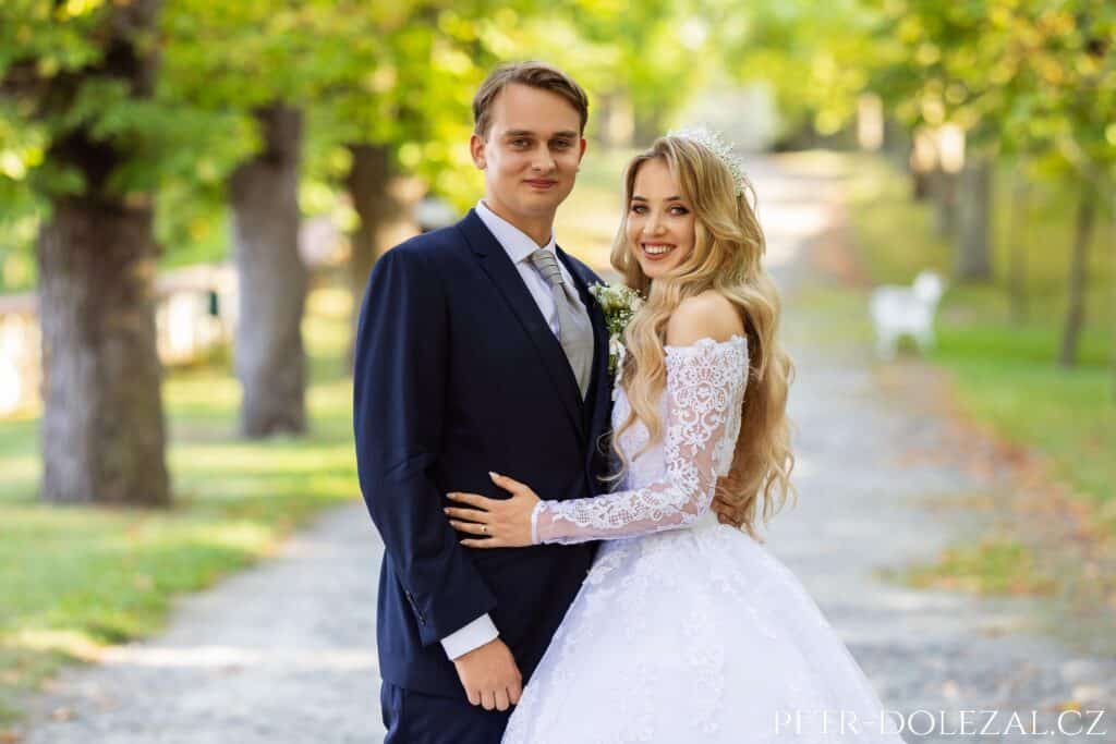 Nevěsta a ženich stojí těly otočení směrem k sobě, dívají se ale do fotoaparátu, nevěsta má položenou ruku na břichu ženicha.