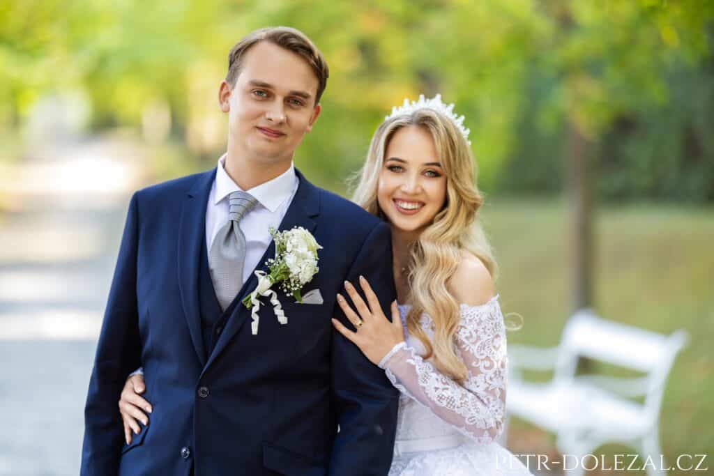 Dvojportrét novomanželů, od pasu nahoru, dívají se do fotoaparátu, nevěsta stojí částečně skrytá za zády ženicha.