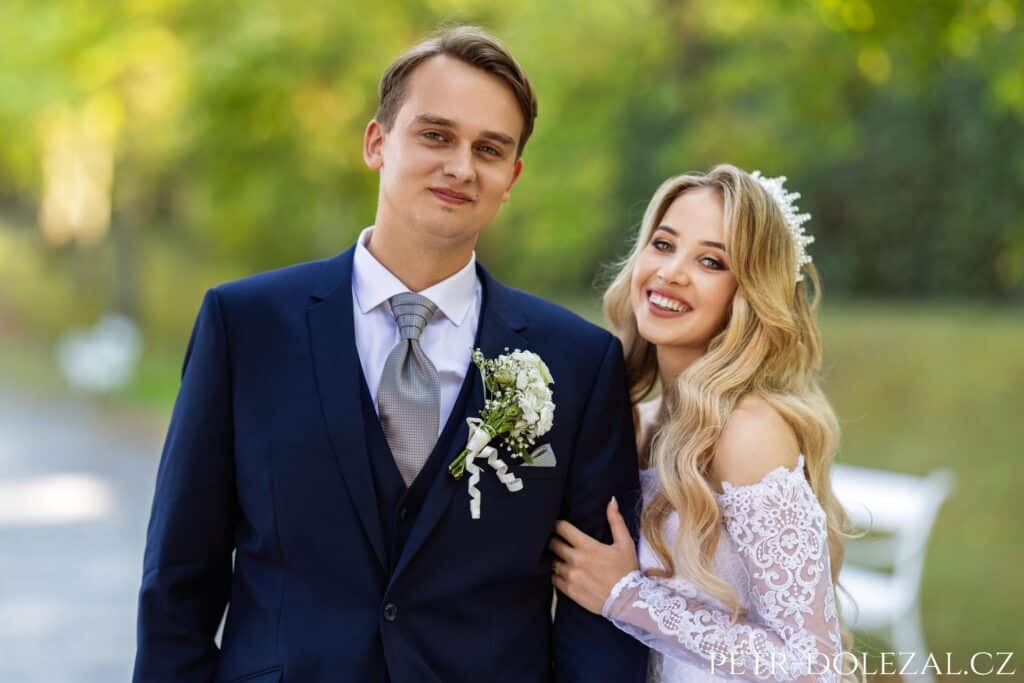 Dvojportrét novomanželů, od pasu nahoru, dívají se do fotoaparátu, nevěsta drží ženicha za paži.