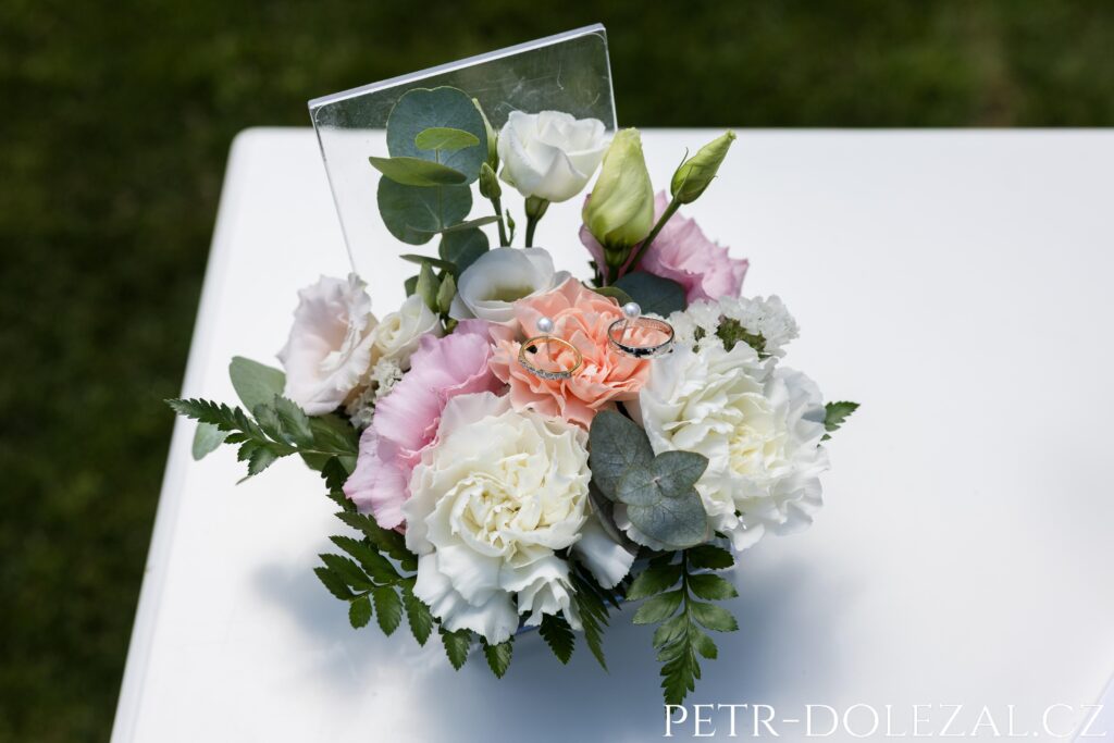 skleněná krabička s květinami a snubními prsteny položená na oddacím stolku na začátku svatebního obřadu