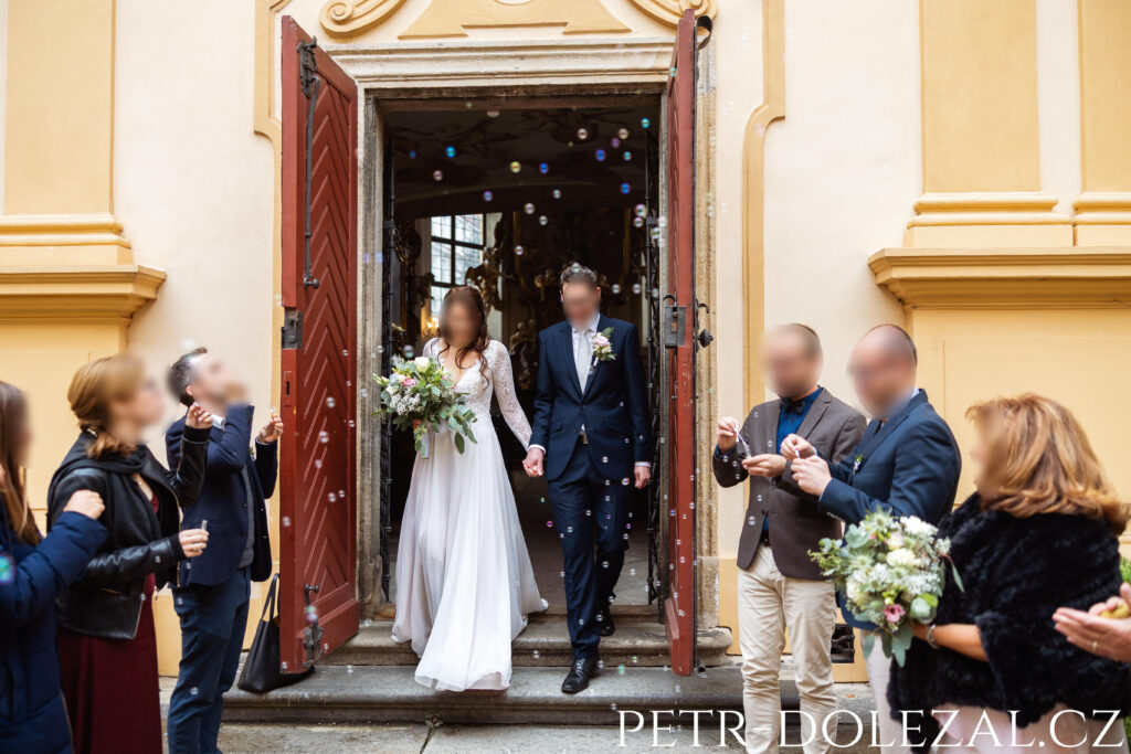 Novomanželé odcházejí špalírem ven z kaple, ve vzduchu bubliny z bublifuků
